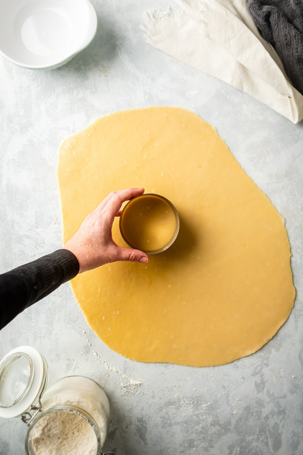 A thin piece of empanada dough flat on a gray counter. There is a hand holding a round cutter on top of the middle of the sheet of dough.