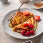 White plate with a slice of French toast that is cut in half on top. There are some raspberries on the plate and a slice of strawberry on each piece of French toast with a fork on the edge of the one slice.