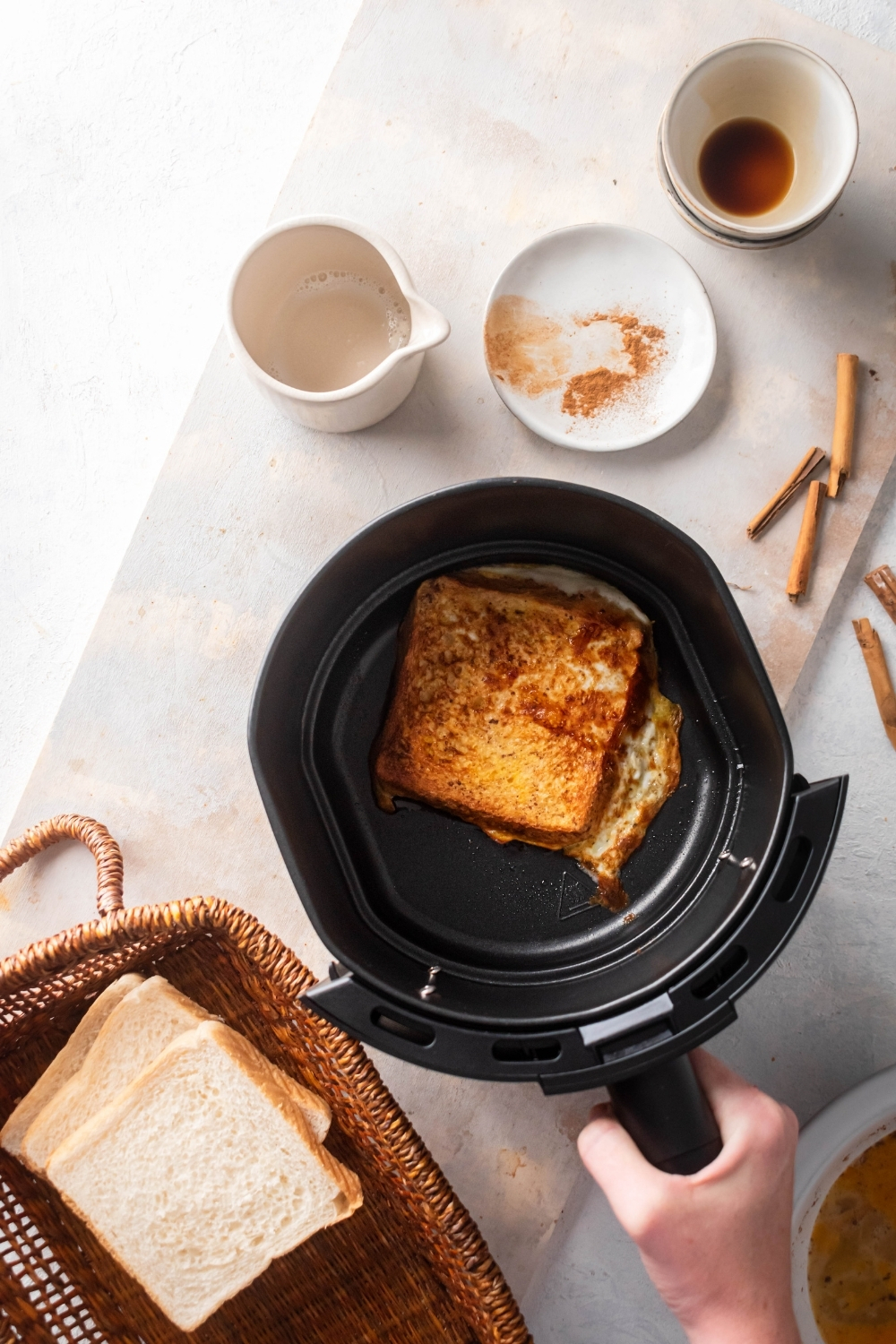 Part of the basket with three slices of bread stacked on top of each other, a piece of French toast in an air fryer, white picture, a plate with some cinnamon on it, and a white bowl on a white counter.