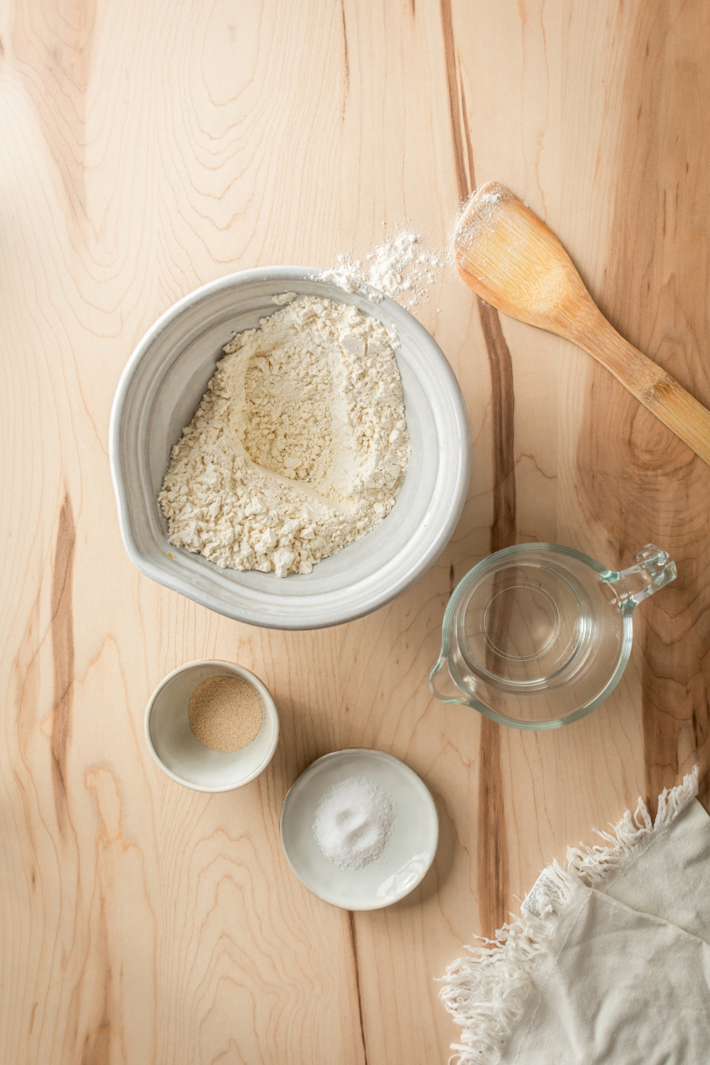 A large white bowl filled with flour, a small bowl filled with active yeast, a small dish filled with salt, a small pitcher filled with water, and a wooden spoon all on a wood counter.