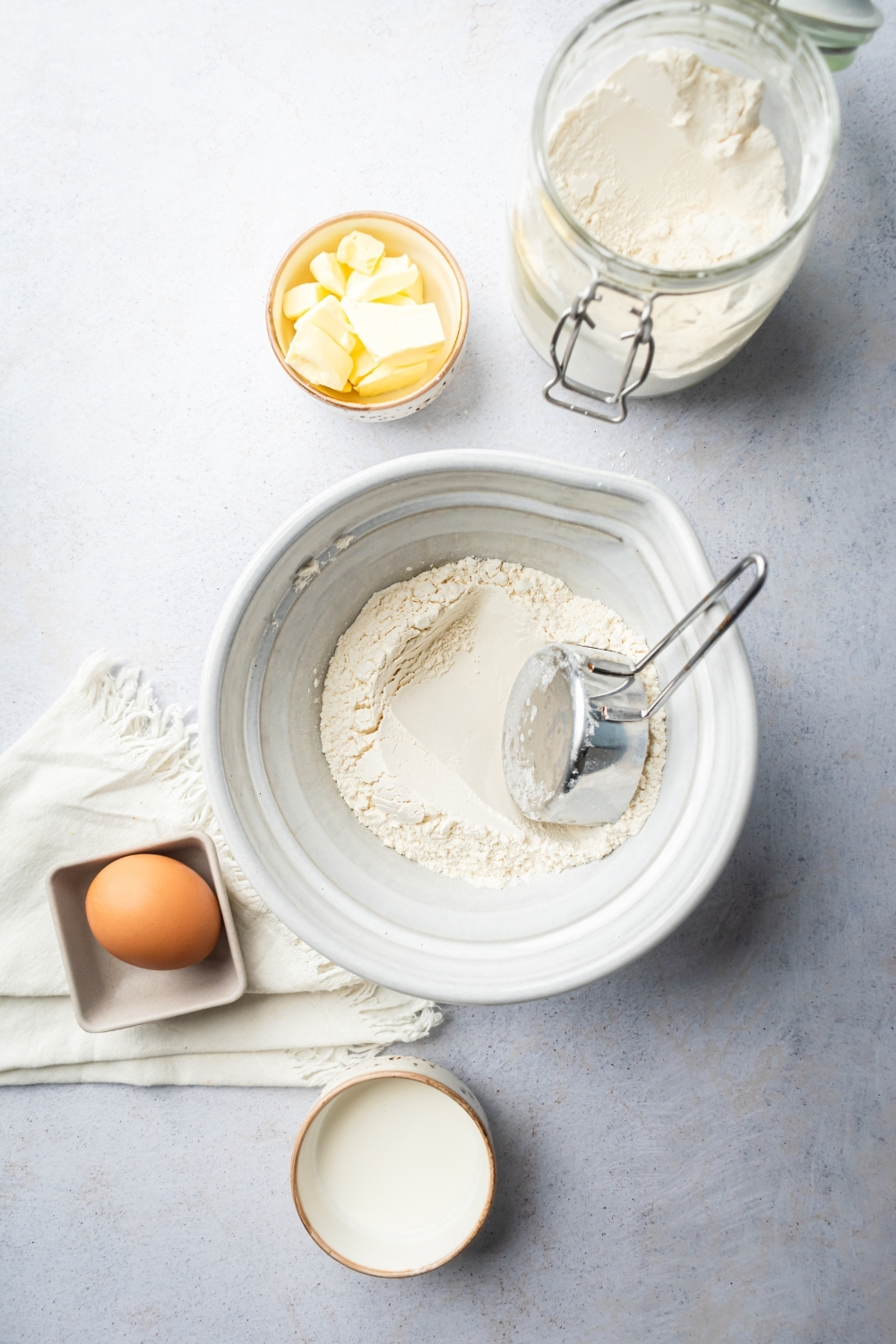 A white bowl filled with flour, an egg, small bowl with butter cubes, and a glass jar of flour all on a gray counter.