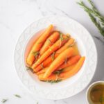 A bunch of carrots with rosemary sprigs on top in a white bowl on the white counter.