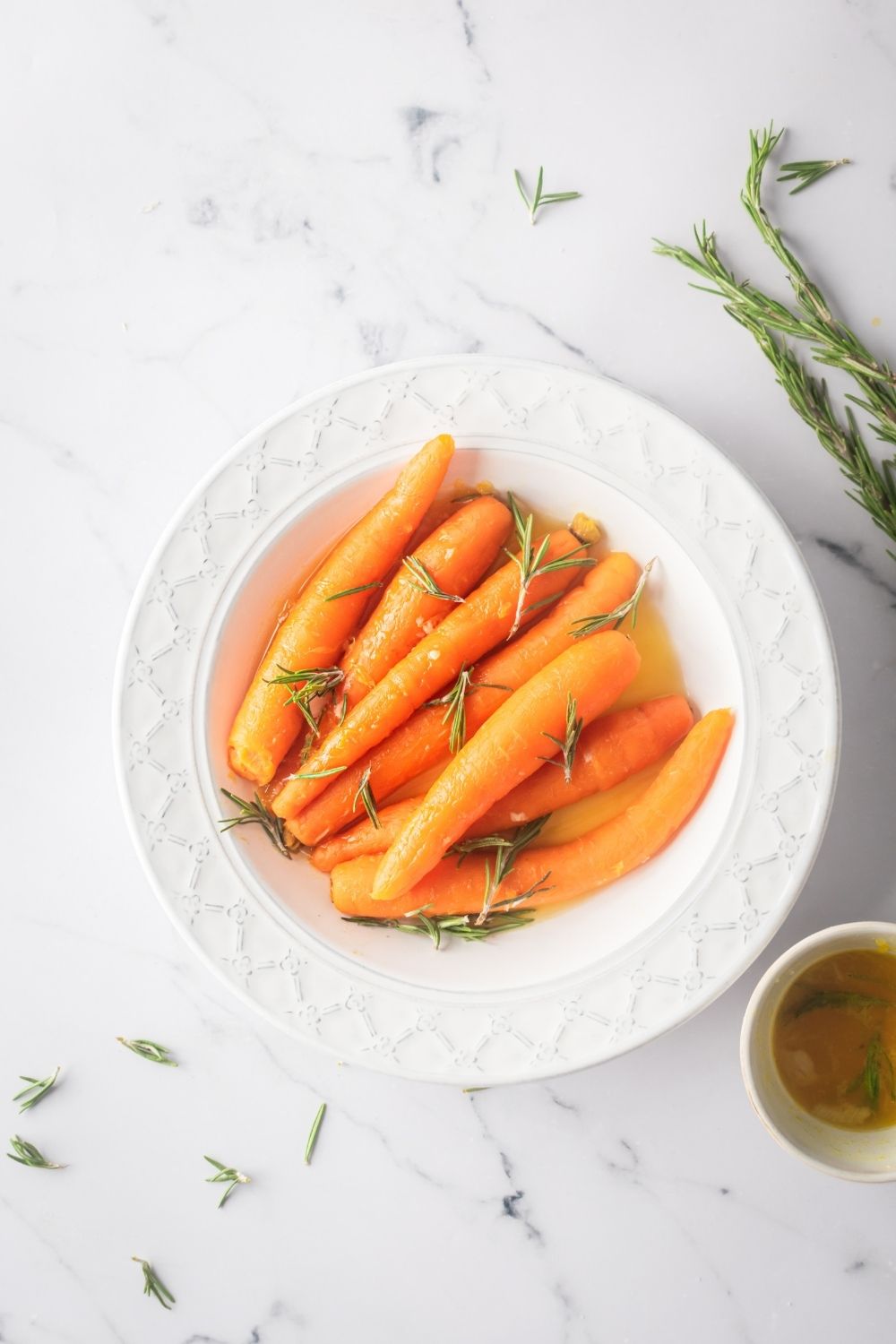 A bunch of carrots with rosemary sprigs on top in a white bowl on the white counter.