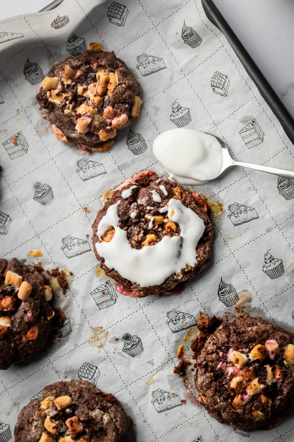 A roll of three chocolate marshmallow cookies on a piece of parchment paper on a baking tray. The cookie in the middle has fluff on top and there is a spoon with fluff on it next to the cookie.