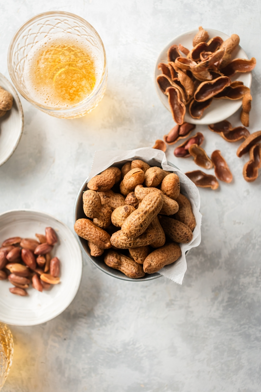 I shelled peanuts in a cup with parchment paper on a gray counter. To the left of that is a small plate of peanuts and to the right a small plate of shells. Behind the cup of shelled peanuts is a glass of beer.