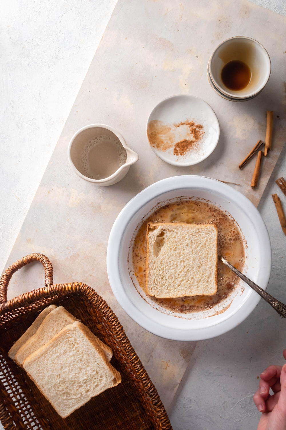 Heard of a basket or three slices of white bread stacked on top each other, a white bowl with French toast mixture and a slice of bread in it, an empty picture, and empty white plate, and an empty bowl on the white counter.