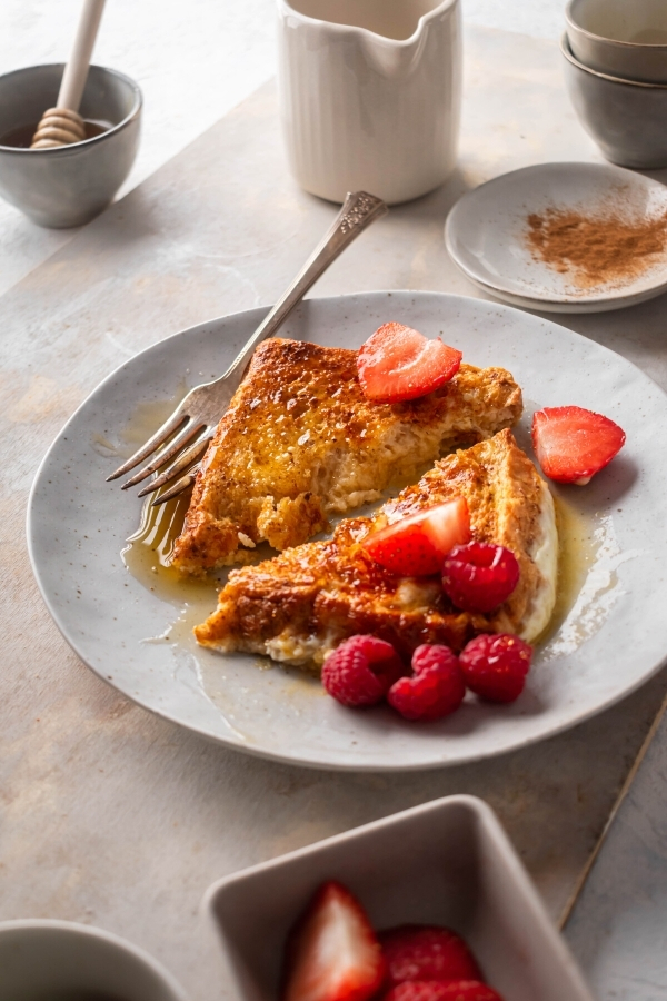 A whiter plate with a piece of french toast sliced in half on it. A fork is on the edge of the one slice and there are some sliced strawberries and raspberries on the plate.