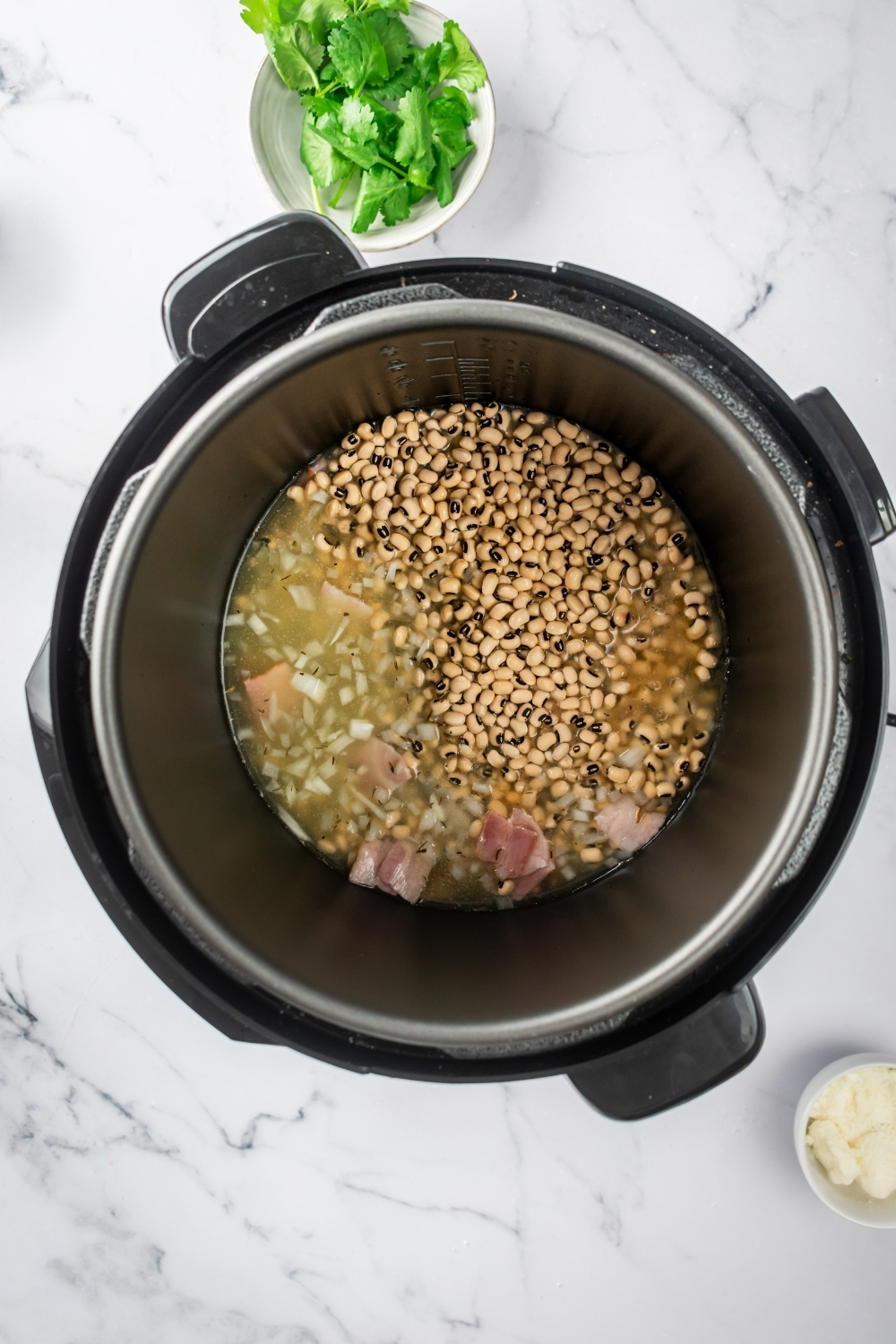 And instant pot on a white counter filled with black-eyed pea's, bacon, onions, and chicken stock.