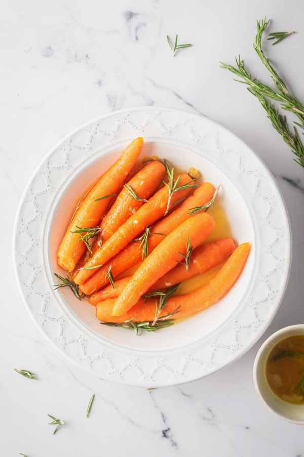 A bunch of carrots with a honey glaze with rosemary sprigs on top in a white bowl on a white counter.