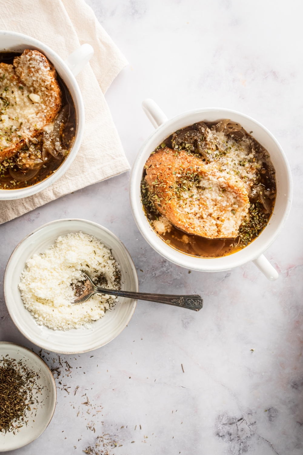 I am white counter with a small white bowl with Parmesan cheese and a spoon in it, a white bowl filled with French onion soup, and a part of a white bowl on top of a white napkin.