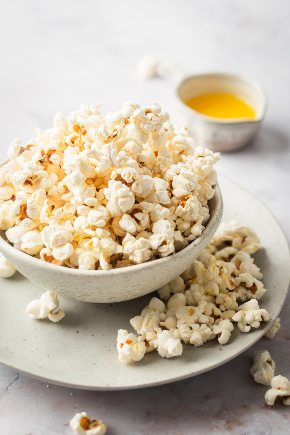A white plate with a bowl of popcorn on it. There are a few pieces of popcorn on the plate and behind is a small white cup of melted butter.