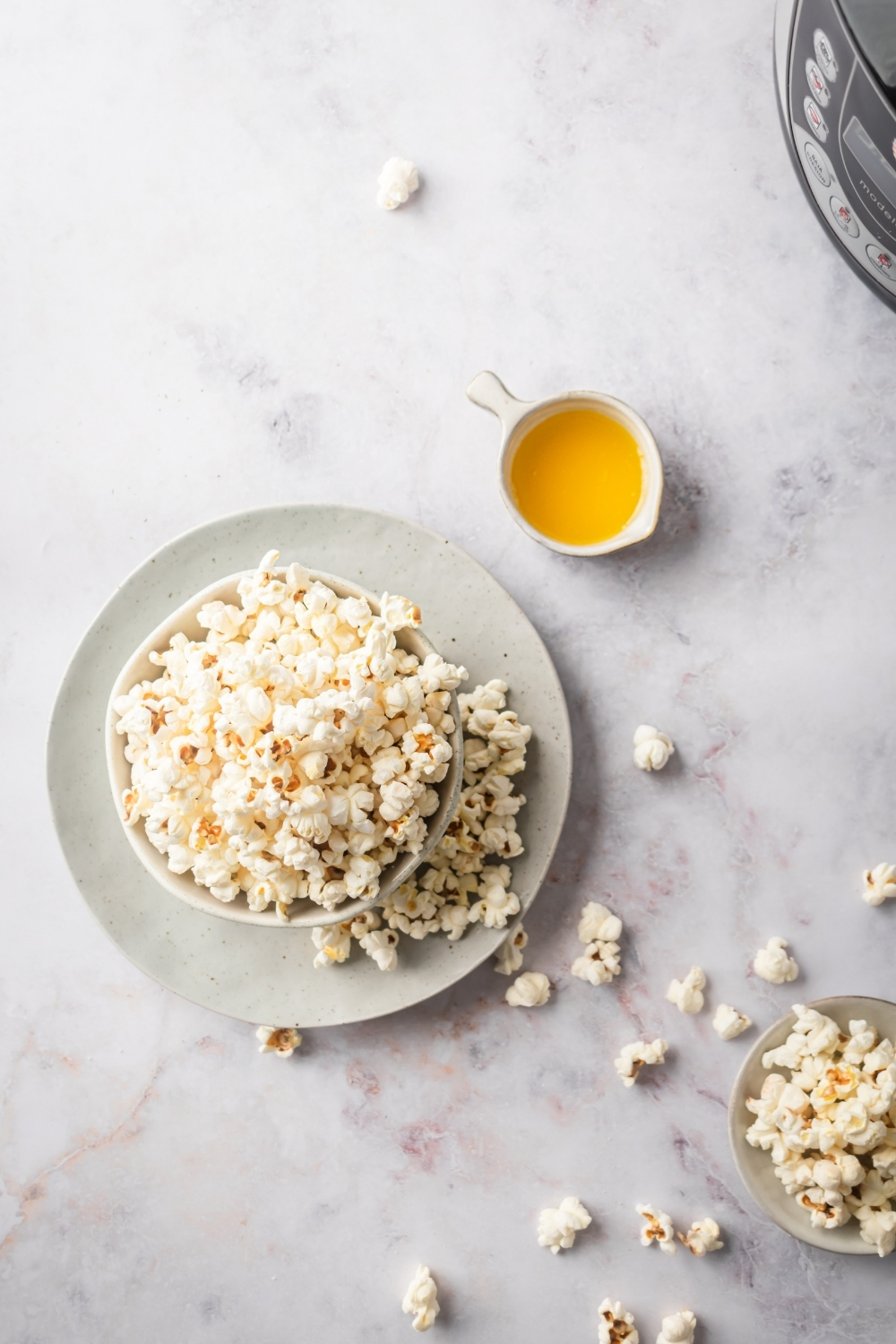 Popcorn in a white bowl on top of a dish and a white counter. Behind it is a small white cup of butter and in front of it are pieces of popcorn on the counter and another small bowl of popcorn.