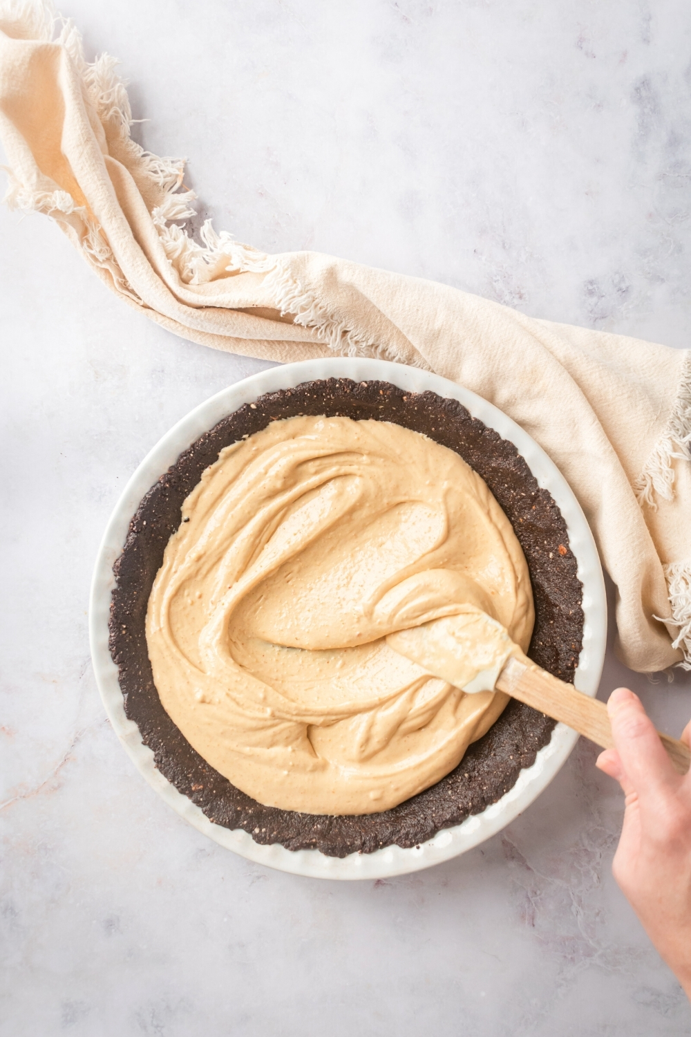 A white pie dish a chocolate almond flour pie crust with peanut butter filling being swirled on top of it with a spatula held by a hand.