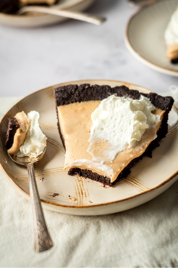 A slice of peanut butter chocolate pie on a plate. A spoon is next to the slice of pie on the plate with a piece of the front of the pie on the spoon.