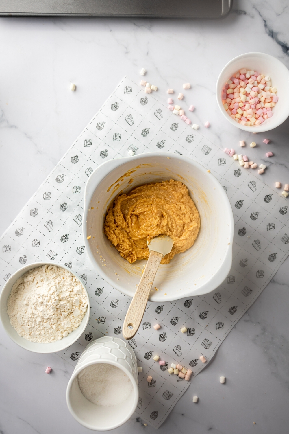 A white mixing bowl with cookie dough for chocolate marshmallow cookies in it. There is a rubber spatula submerged in the door and behind it is a small white bowl flower in front of a small white bowl of mini marshmallows.