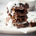 For chocolate marshmallow cookies stacked on top of one another on a piece of parchment paper on a white plate. There is fluff on the top cookie dripping down the front side of it.