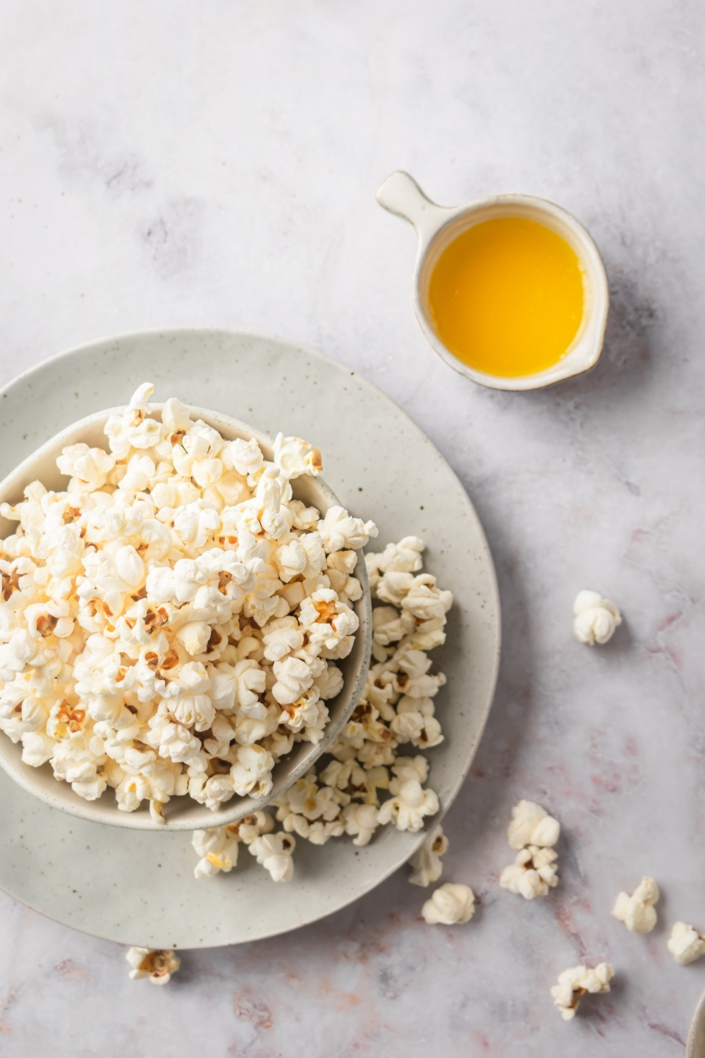 A white bowl filled with popcorn on a dish with a few pieces of popcorn on it on the white counter. Behind the dish is a small white bowl of melted butter.
