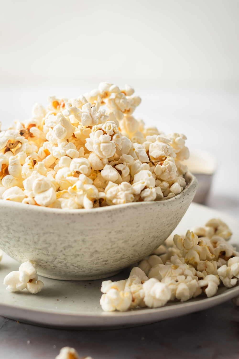 A white bowl filled with popcorn on top of a plate with a few pieces of popcorn on it.