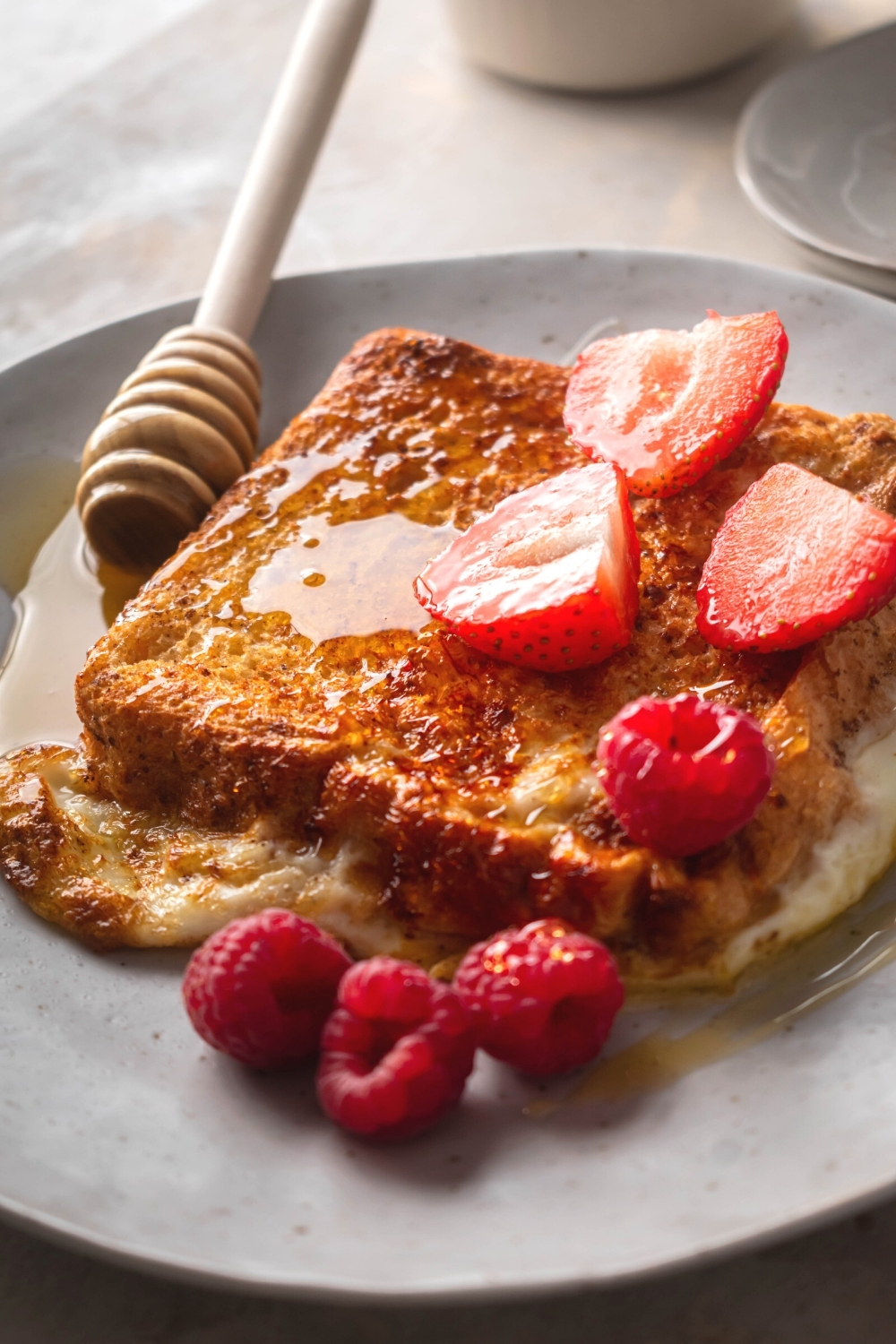 A white plate with a slice of French toast covered and maple syrup with your sliced strawberries and some raspberries.