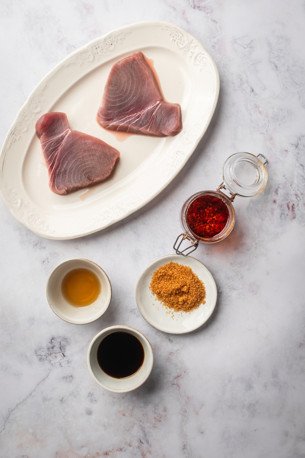 White plate with two tuna steaks on it, a bowl of sesame oil, a bowl of soy sauce, a small white plate with brown sugar on it, and a glass jar with chili flakes in it on a grey counter.
