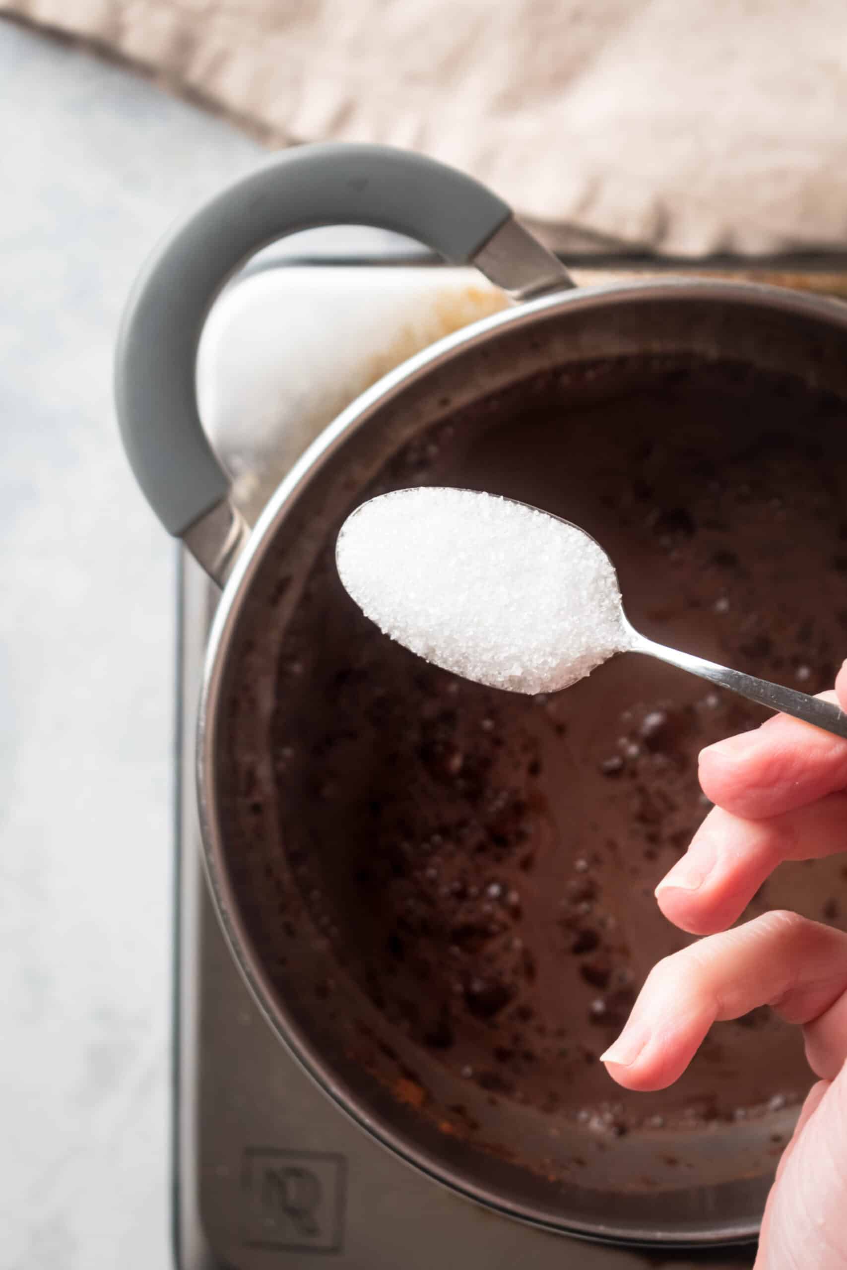 A hand holding a spoon with granulated erythritol over a pot filled with hot chocolate.