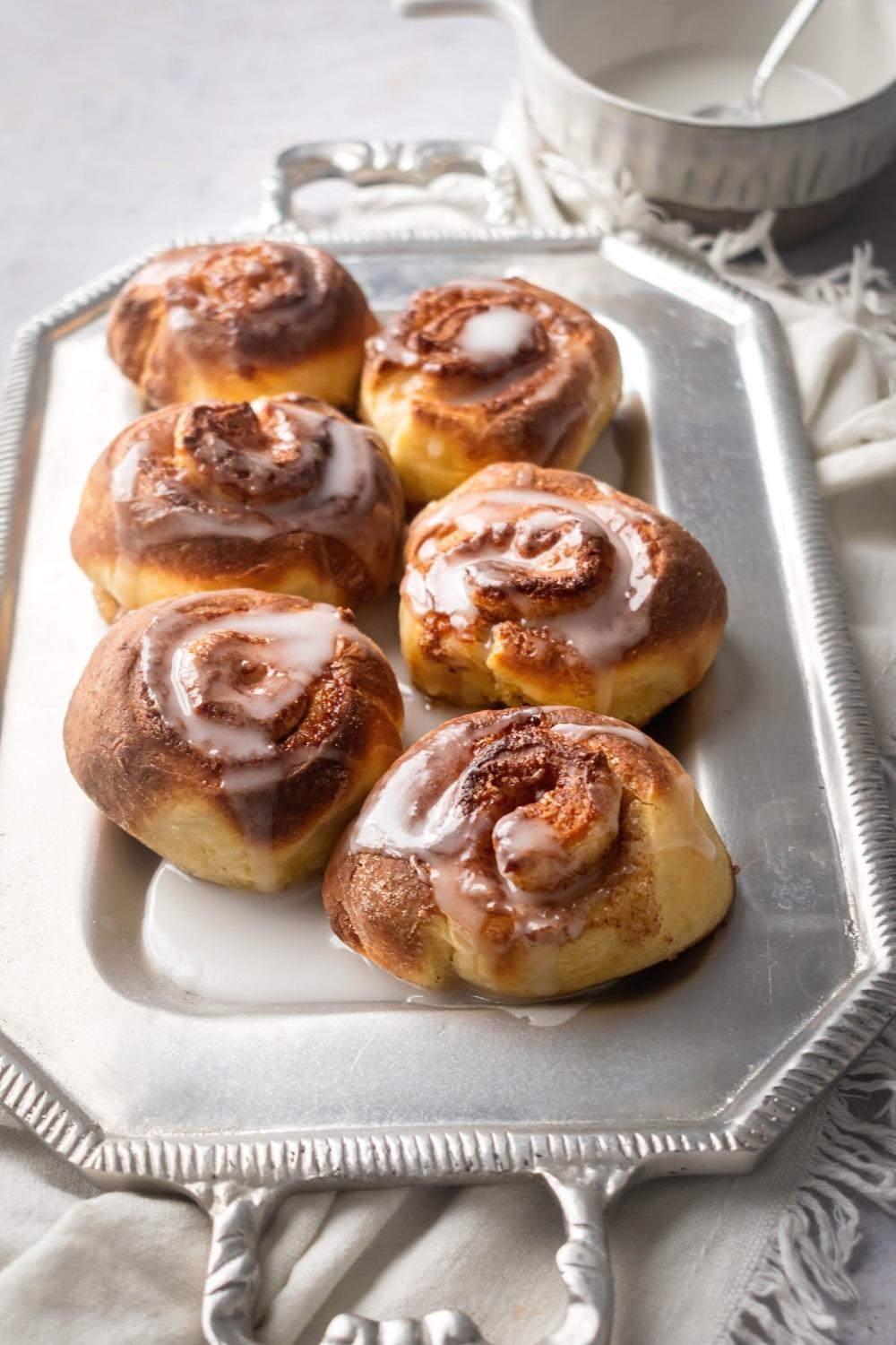 Six cinnamon rolls in two rows of three on a rectangle silver serving dish. Behind it as part of a small white bowl of icing.