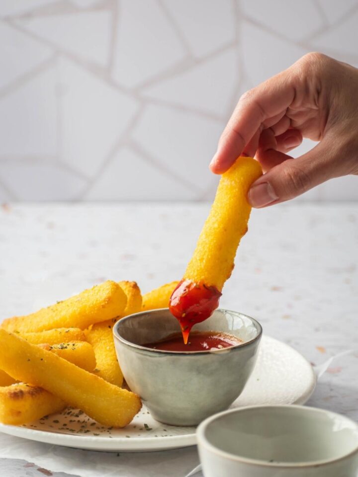 A hand holding a mozzarella stick dipping it in a cup of marinara sauce. The marinara sauce is next to some other mozzarella sticks.
