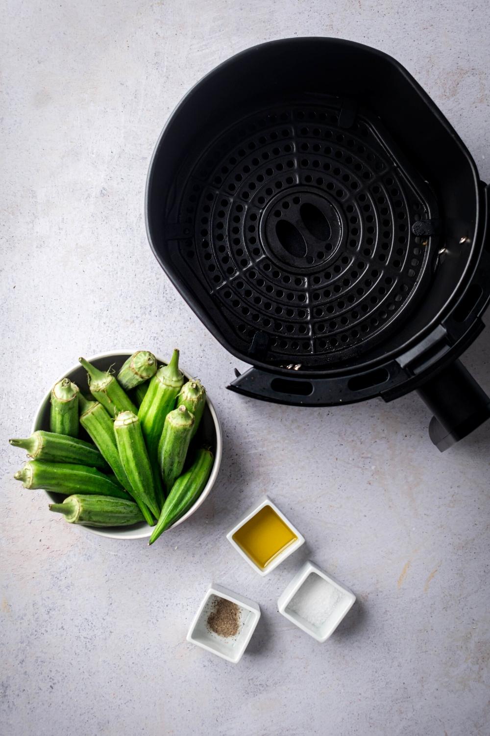An air fryer, a bowl filled with okra, a small cup of olive oil, a small cup of salt, and a small cup of pepper all on a grey counter.