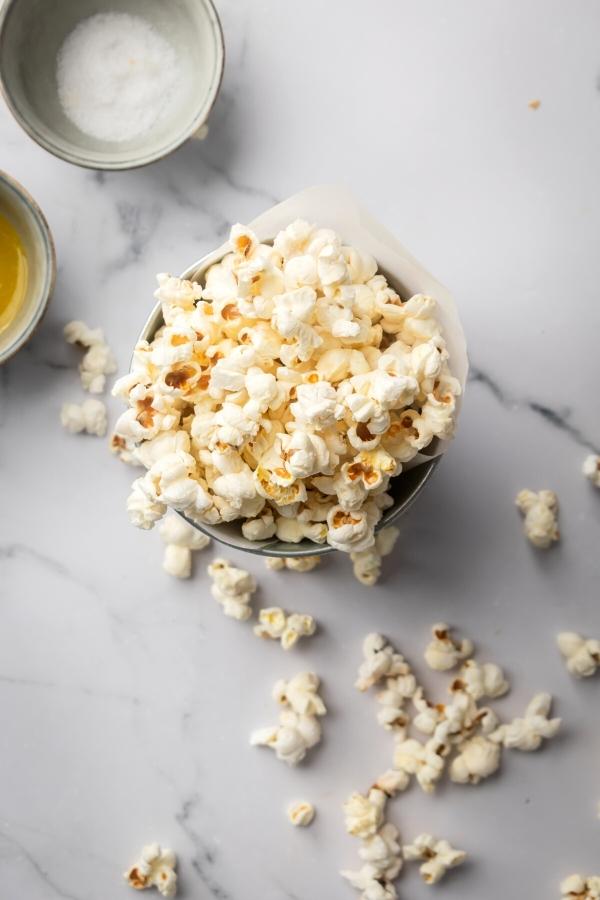 Popcorn in a tin cup on top of a grey counter. There is some popcorn around the cup and behind it is part of a small bowl of butter and a small bowl of salt.