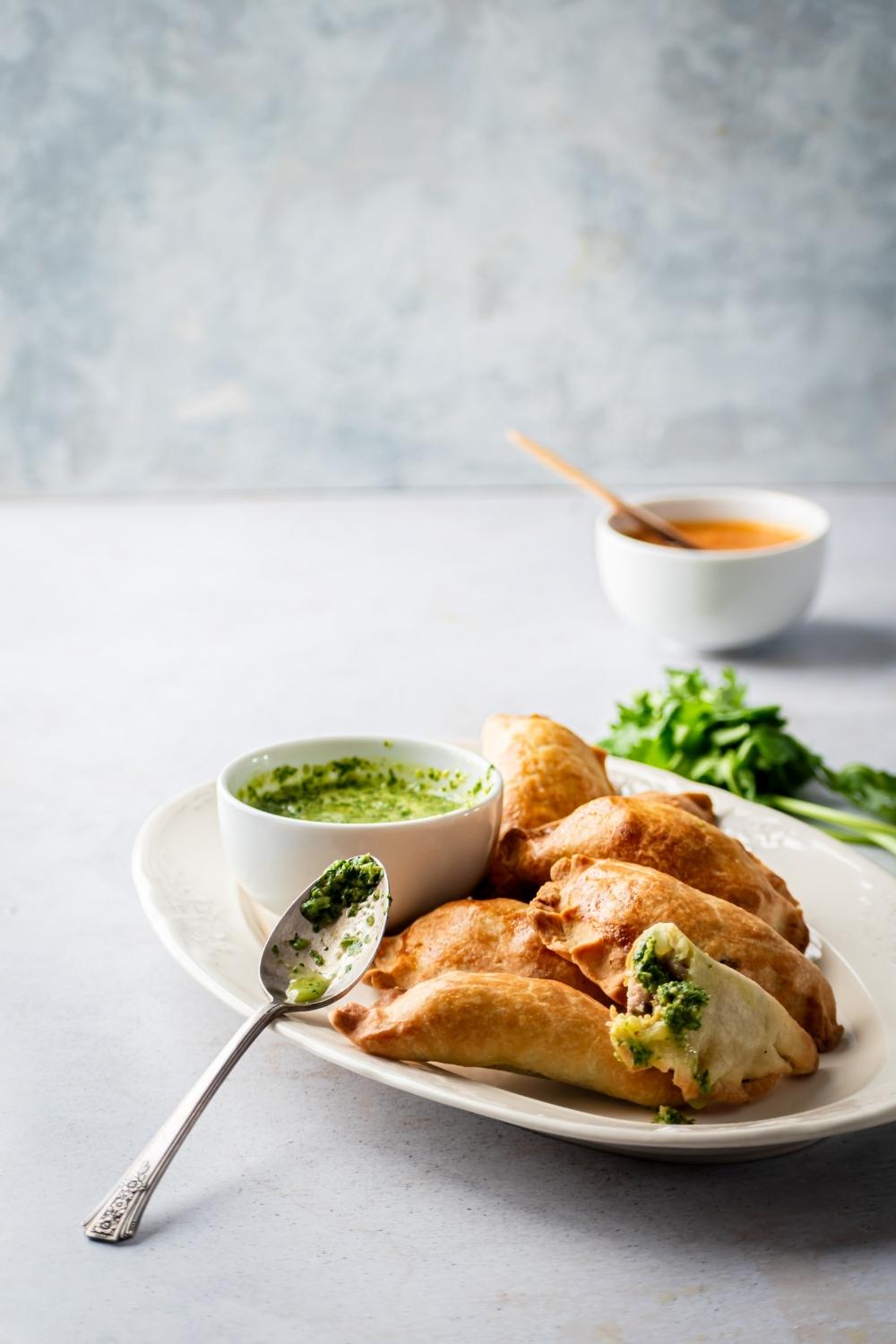 A white oval shaped plate with a bunch of beef empanadas on it and a small white cup of salsa Verde. There is a spoon leaning against the plate with some salsa Verde on it.