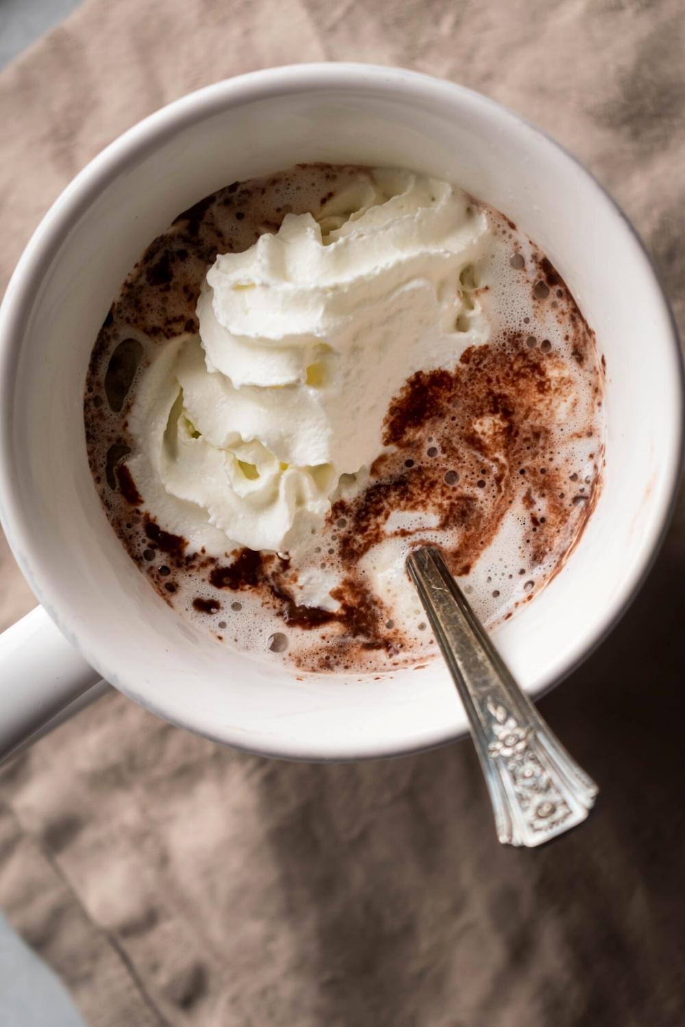 A white mug filled with hot chocolate with whipped cream on top of one side and a spoon submerged in the other side.