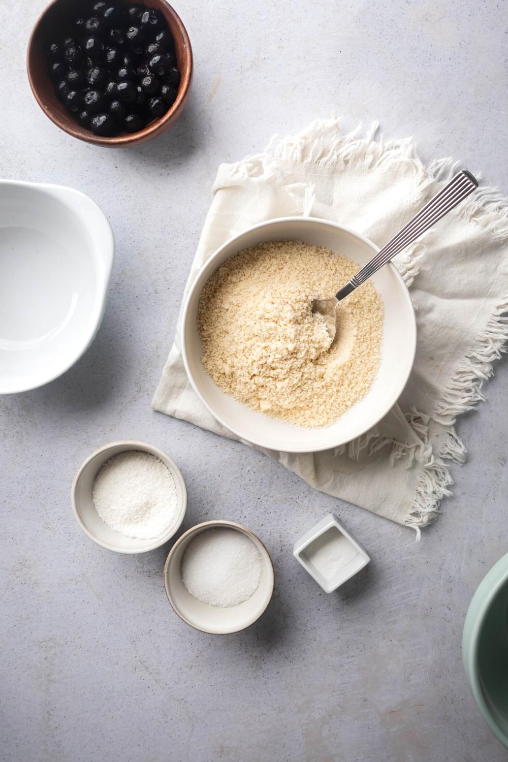 A white bowl with almond flour in it on a white cloth on a grey counter. Behind it on the gray counter is a small bowl of granulated erythritol in front of it a bowl of blueberries.