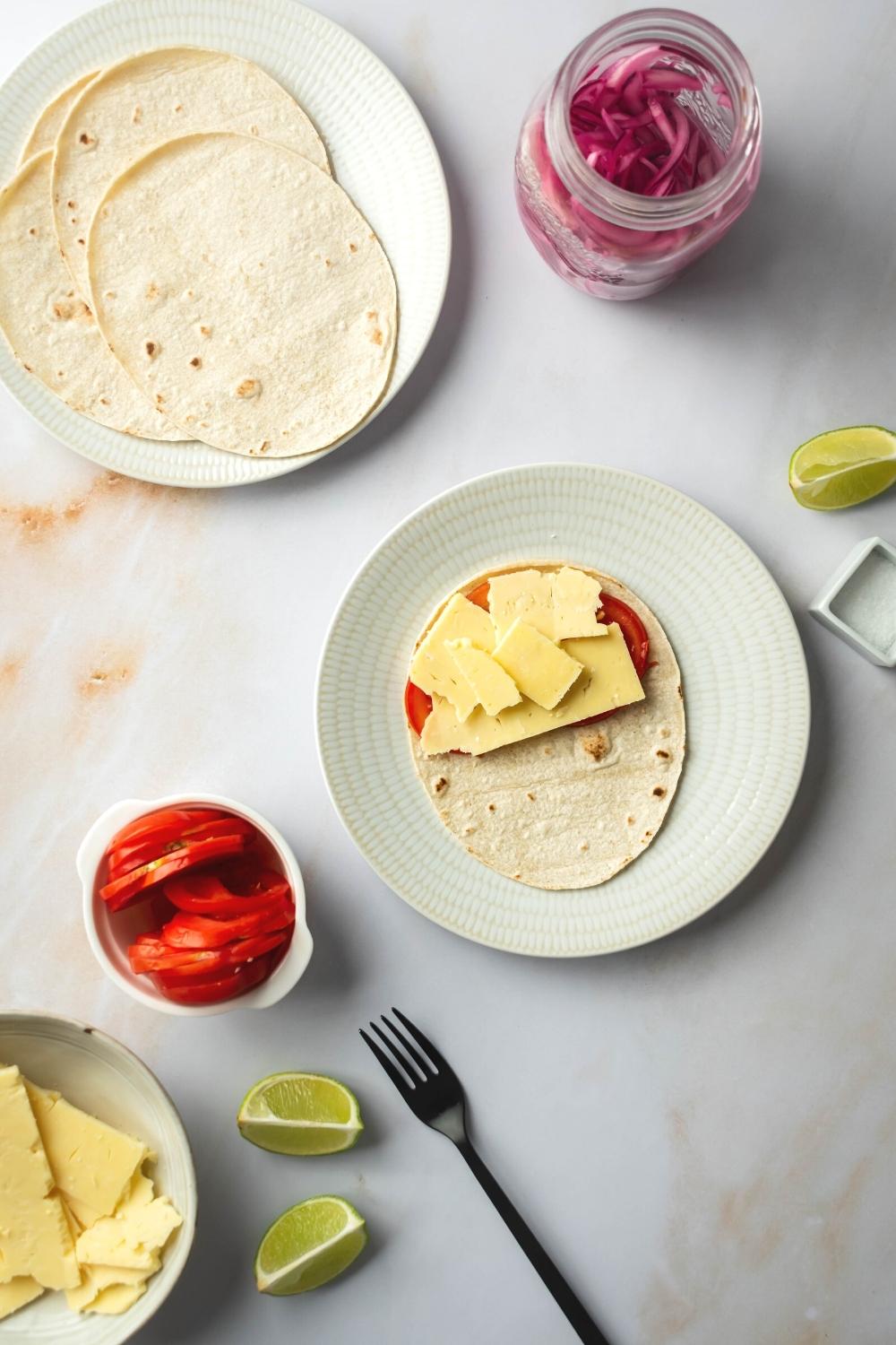 White plate with a tortilla on it with tomato and slices of cheese on top. Next to it is a small cup of sliced tomatoes and behind it as a plate with tortillas on it.