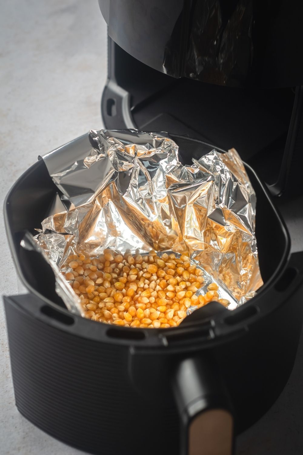 An air fryer basket lined with tin foil with popcorn kernels in it.