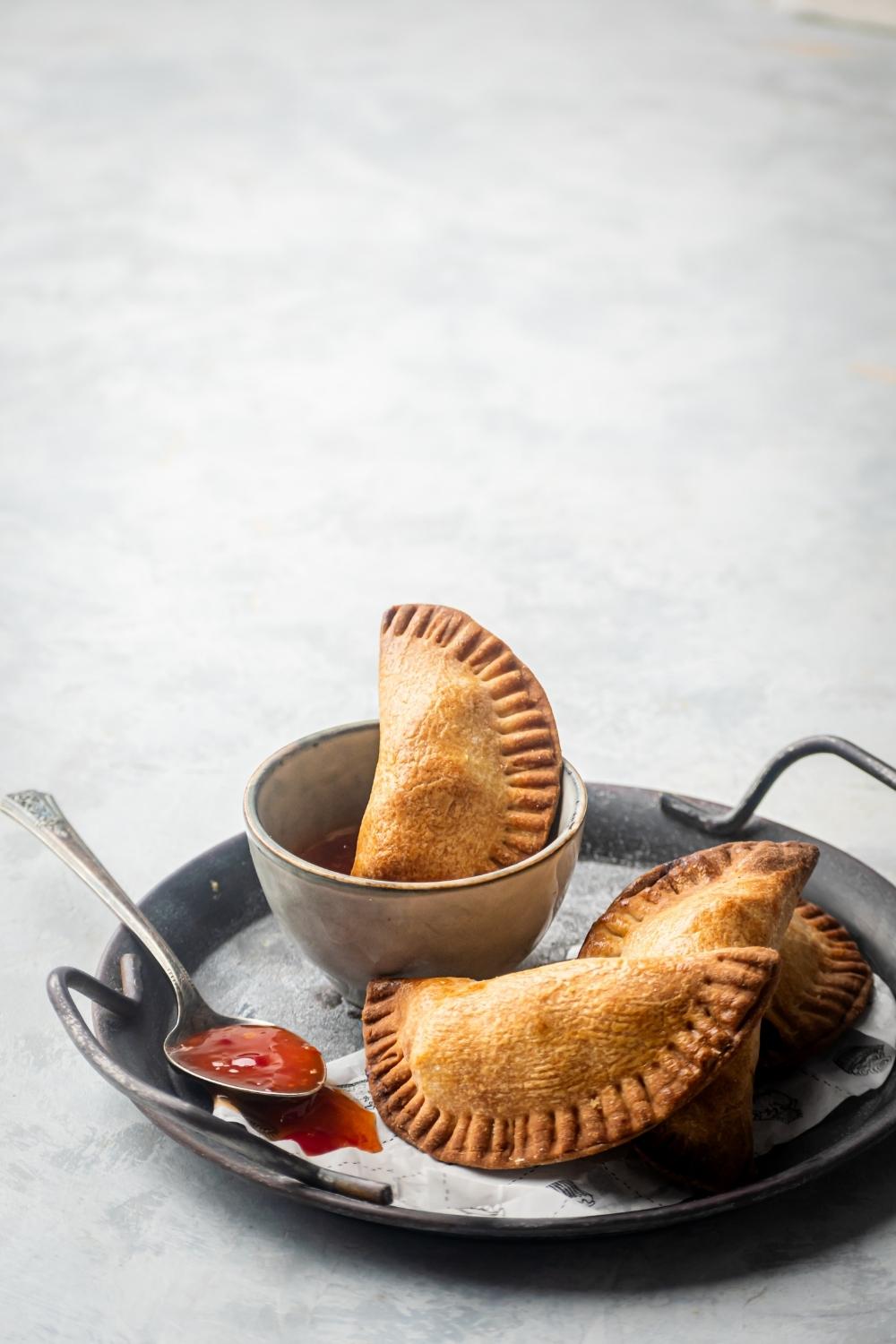 Gray pan with three chicken empanadas on top of one another and one chicken empanada submerged in a small bowl of sweet and sour sauce. There's a spoon on the pan with sweet-and-sour sauce on it and pants and a gray counter.