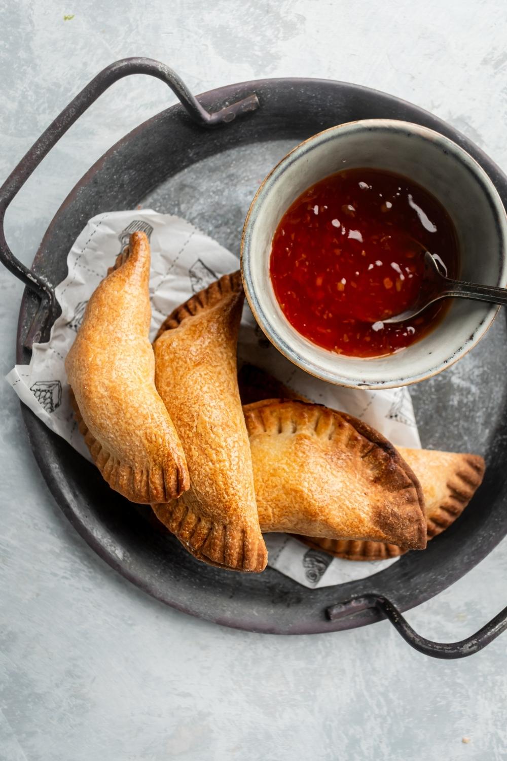 Part of a gray pan lined with parchment paper with four chicken empanadas overlapping one another. There's a small white bowl filled with sweet-and-sour sauce with a spoon in it.