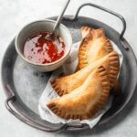 A black pan with four chicken empanadas overlapping one another on it. There is a bowl of sweet and sour sauce on the pan with a spoon in it.