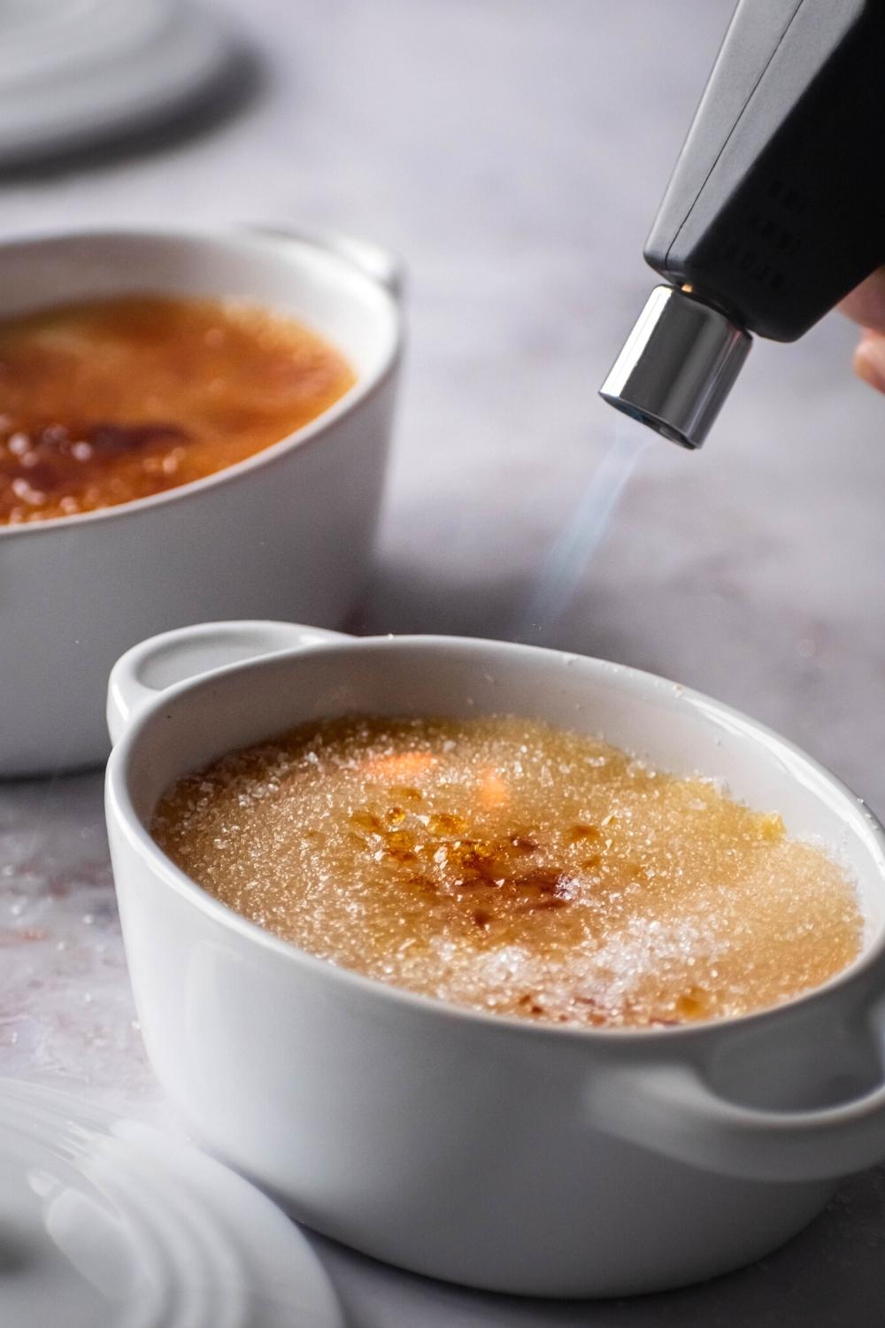 A blow torch torch in the top of crème brûlée in a white ramekin. Behind that is part of another ramekin filled with crème brûlée.