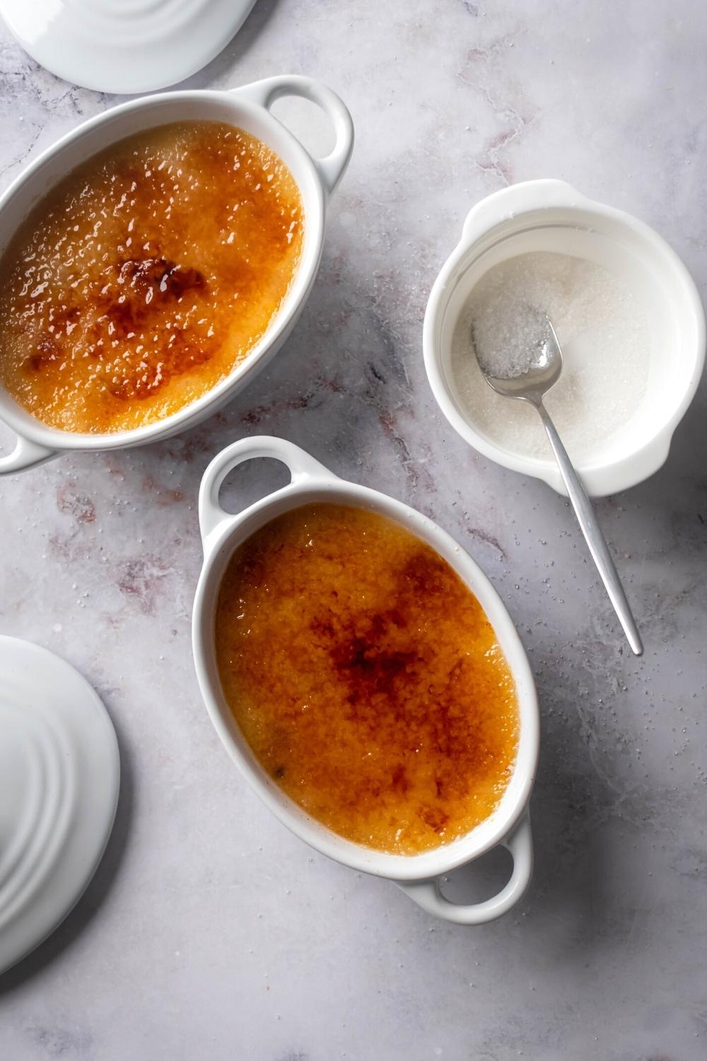 Two large ramekins filled with crème brûlée on top of a white counter. Next to the ramekins to the right is a white bowl filled with sugar with a spoon in it.