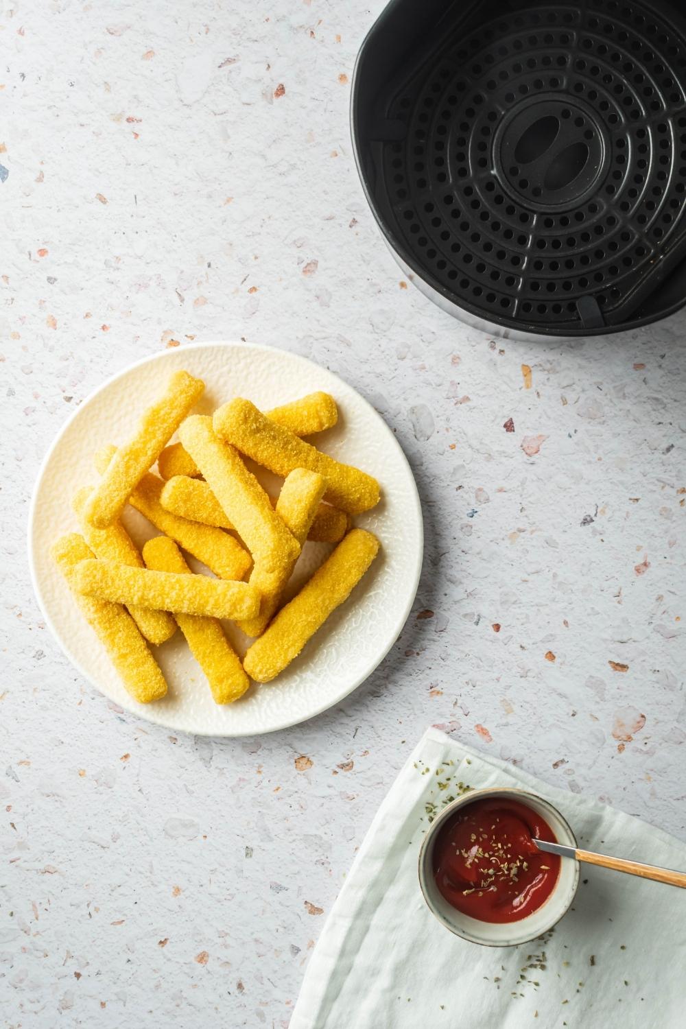 A white plate with a bunch of frozen mozzarella sticks on it. Behind the plate is part of an air fryer.
