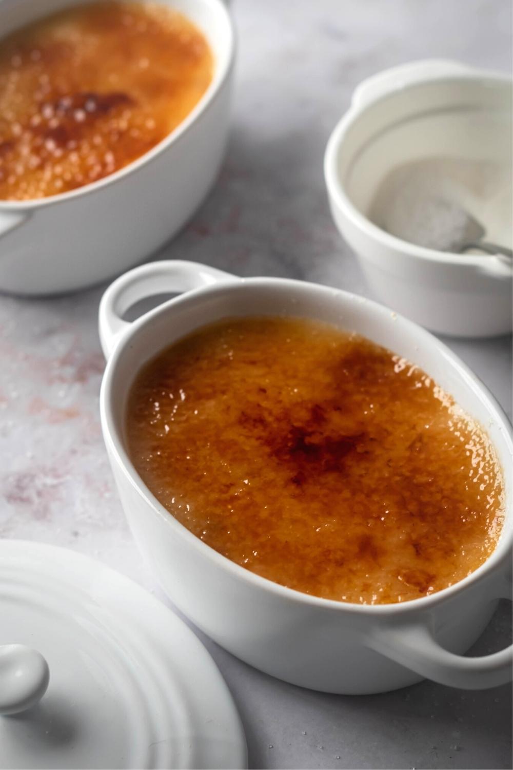 A large white ramekin with crème brûlée in it. Behind it is part of another ramekin with crème brûlée and part of a small white bowl filled with sugar.