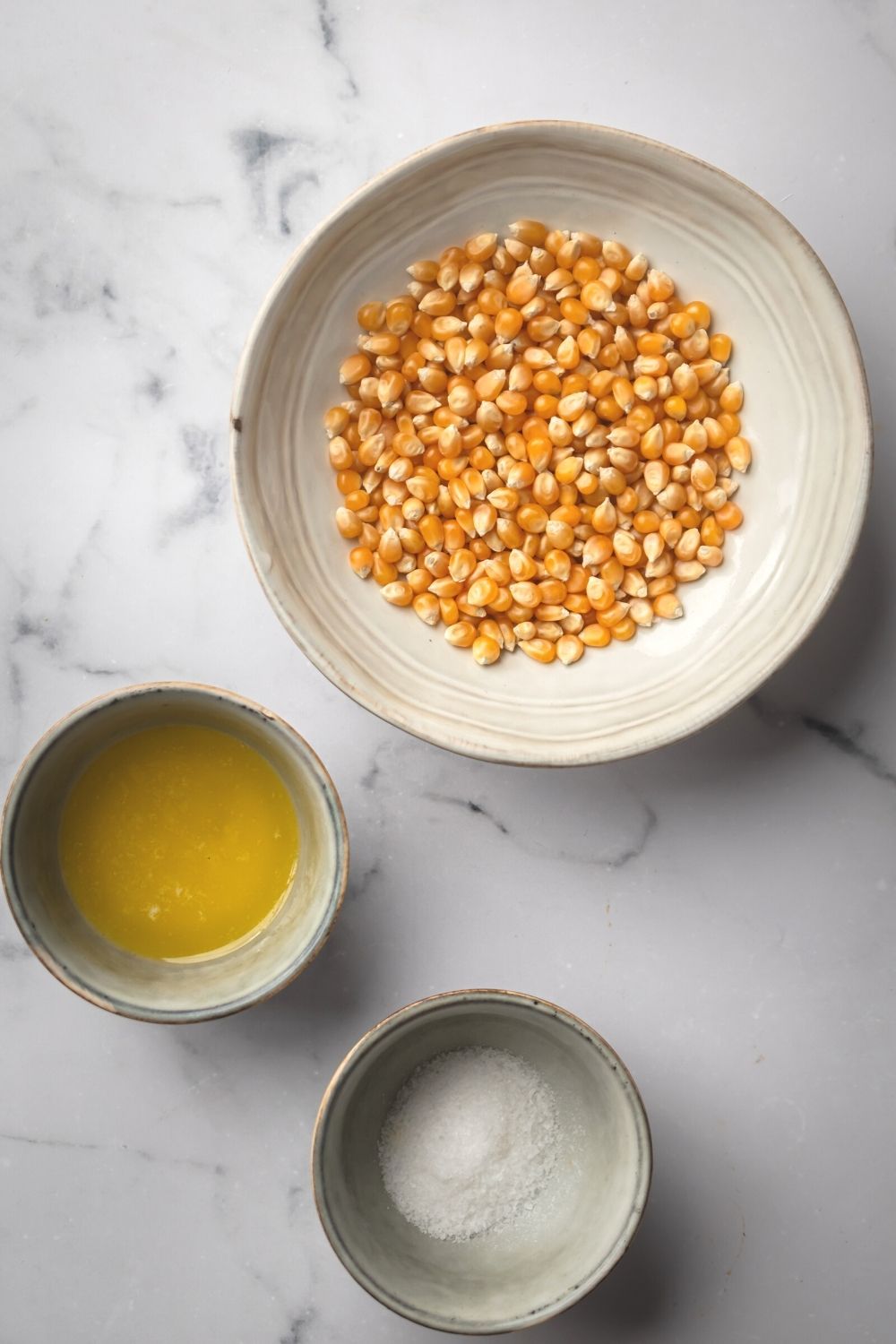 A white bowl of popcorn kernels, a small bowl of butter, and a small bowl of salt on a grey counter.