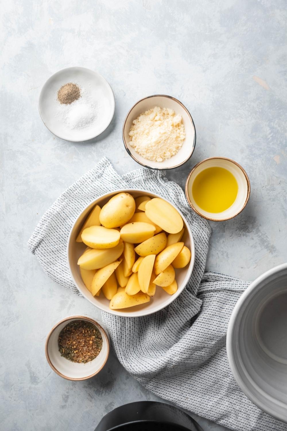 A white bowl with potato wedges in it, a small bowl of olive oil, a bowl of Parmesan cheese, a plate with salt and pepper on it, and a bowl of Italian seasoning all surrounding the bowl of potato wedges on a gray counter.