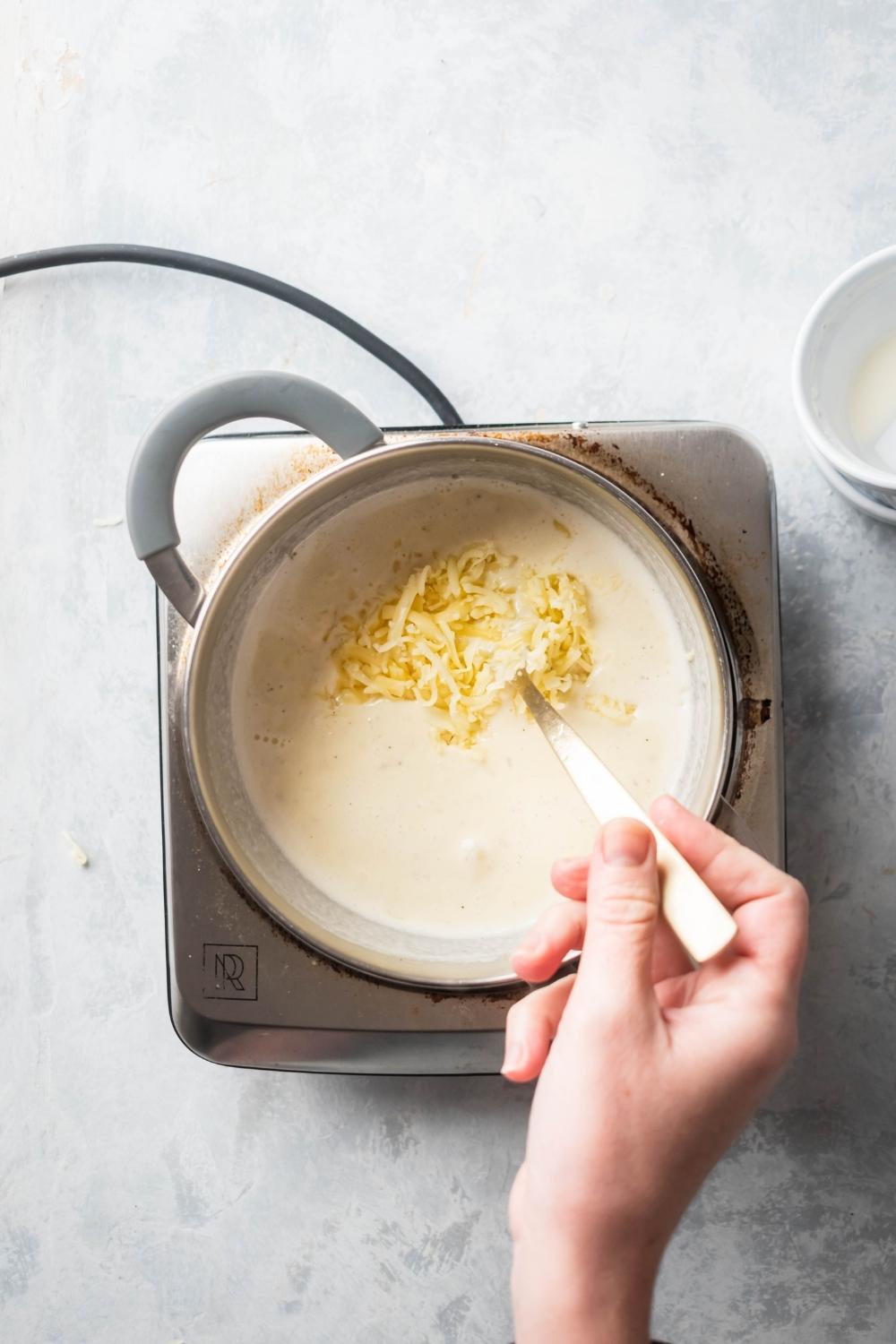 A hand holding a spoon submerged in a pot of cheese sauce. The pot is on top of a burner on a grey counter.