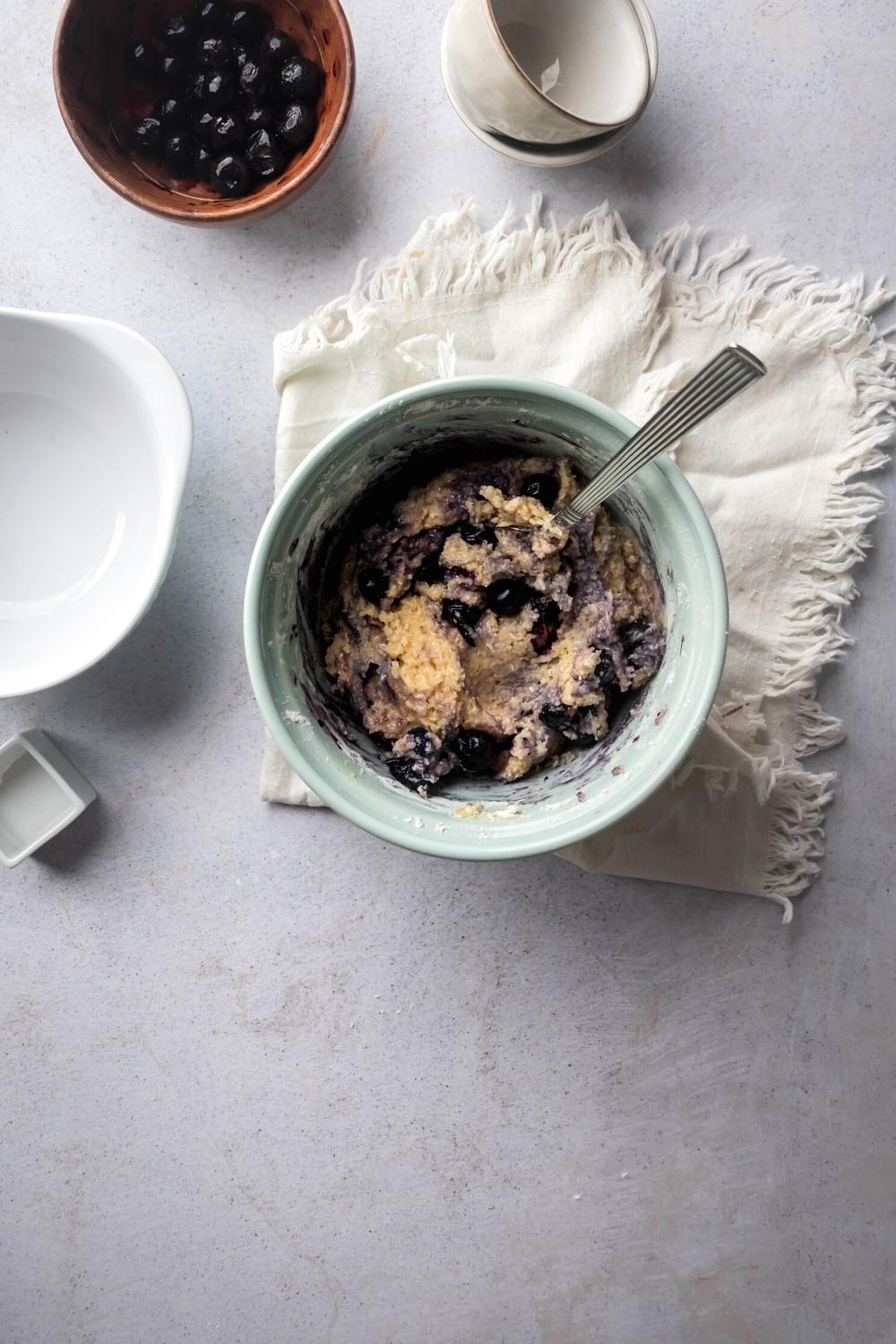 A bowl filled with mixed blueberry scone ingredients. The bowl is on a white cloth on a gray counter and in front of the cloth as a bowl of blueberries and two empty small bowls on top of one another.