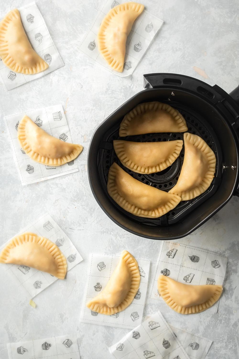 A gray counter with an air fryer filled with four chicken empanadas on it, and six squares of parchment paper with a chicken empanada on top of each surround in the air fryer.