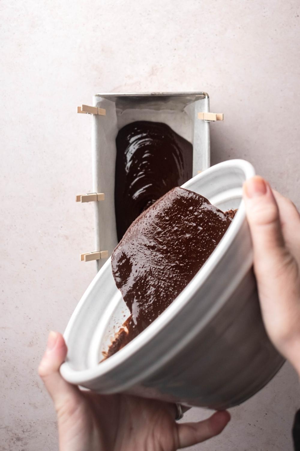 Two hands holding a white bowl with melted chocolate in it, pouring it into a rectangular tin lined with parchment paper.