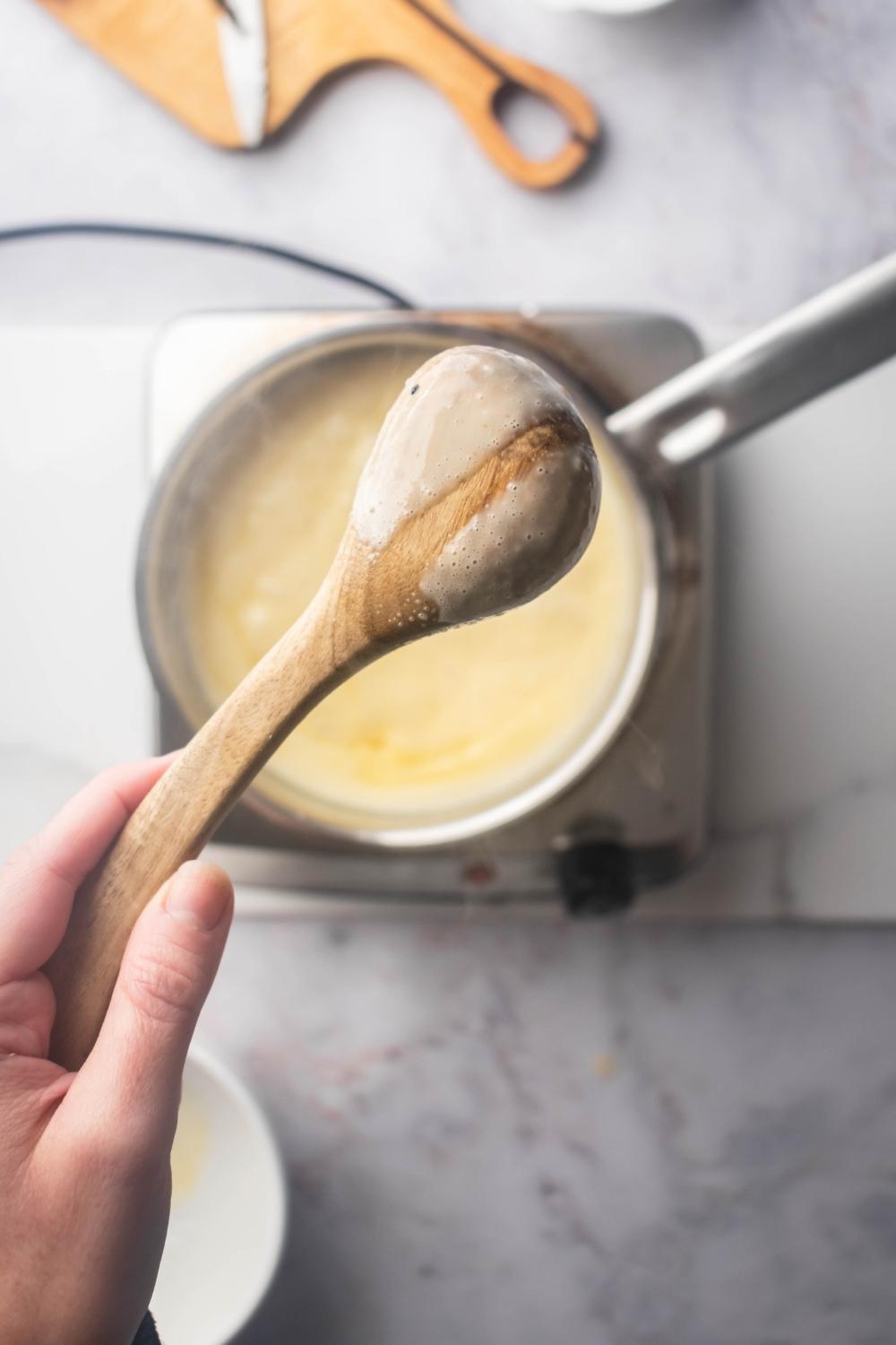 A hand holding a spoon with crème brûlée filling on it with a streak out of the middle. The spoon is being held over a pot filled with crème brûlée on top of a burner on a white counter.