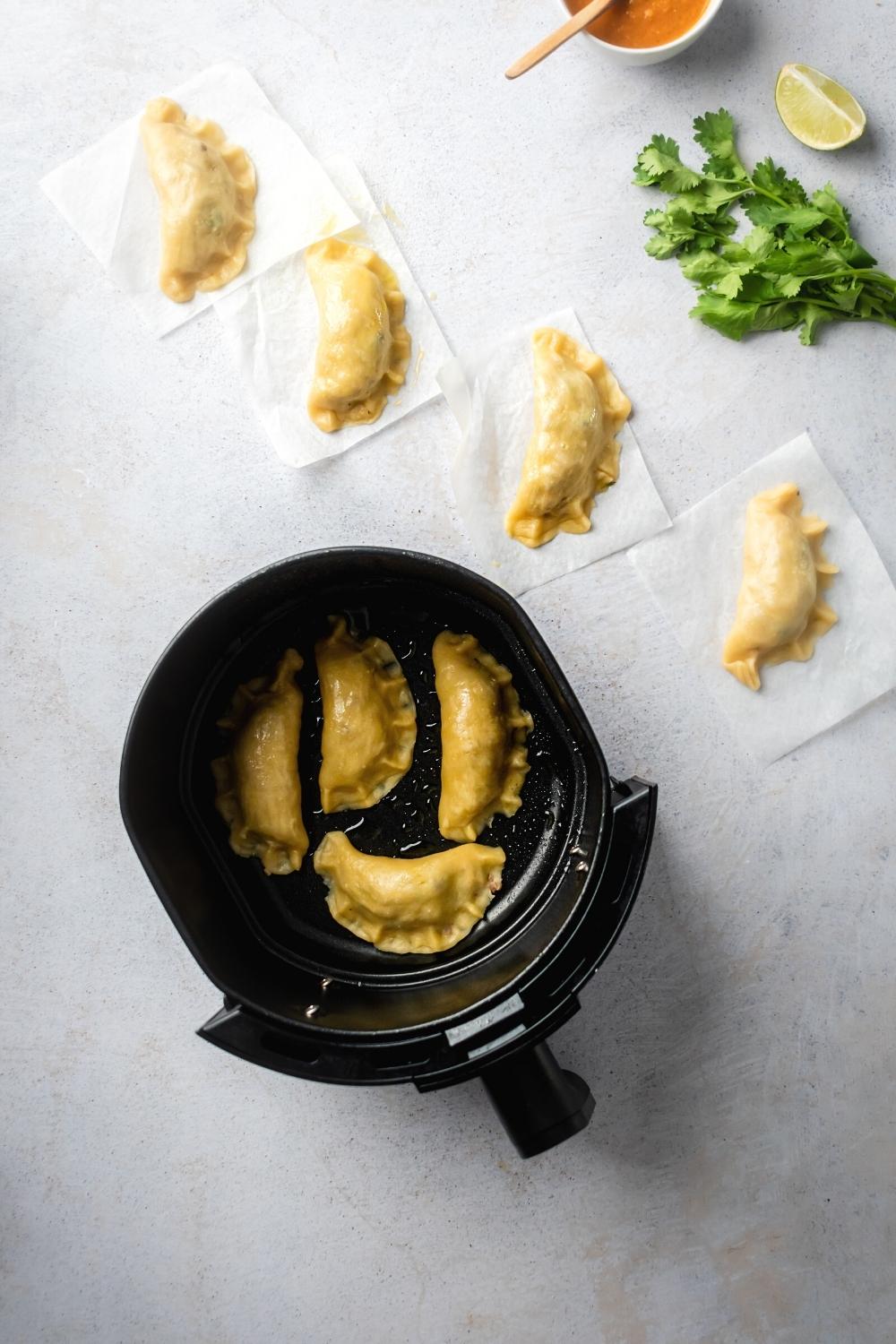 An air fryer filled with four ground beef empanadas and a gray counter. Behind it is for empanadas lined up on squares of parchment paper.