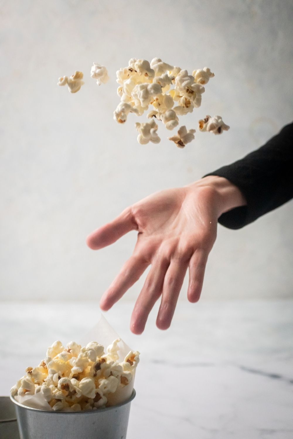 A hand tossing popcorn in the air. Below the hand is part of a tin cup filled with popcorn.